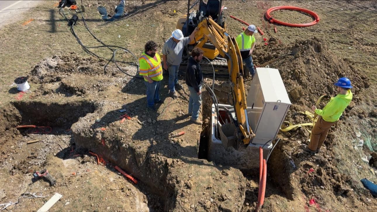 Excavation crew lowering a utility cabinet into a trench with mini excavator